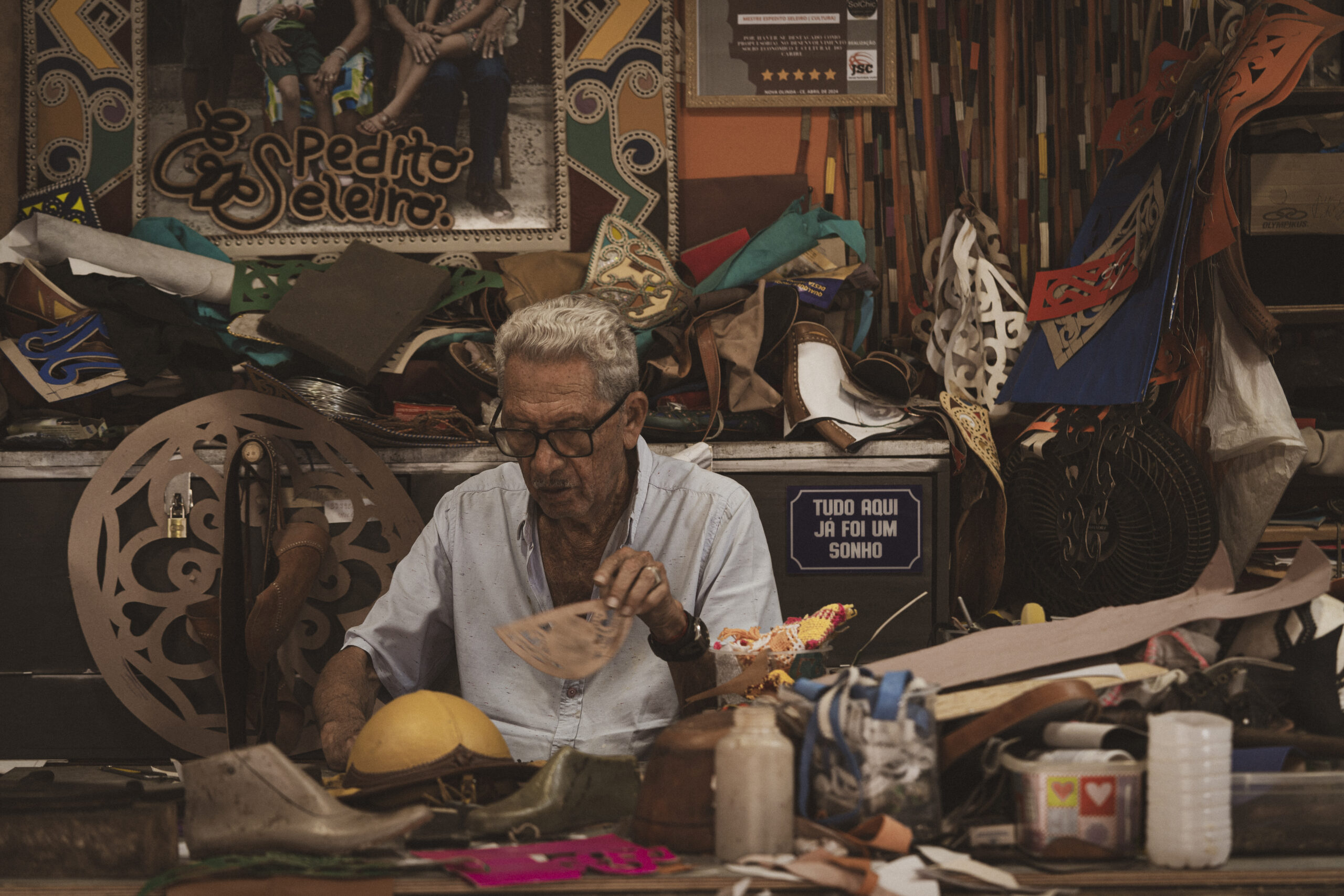 Mestre artesão trabalha uma peça de couro em sua oficina, cercado por materiais, ferramentas e criações em diferentes etapas de produção.