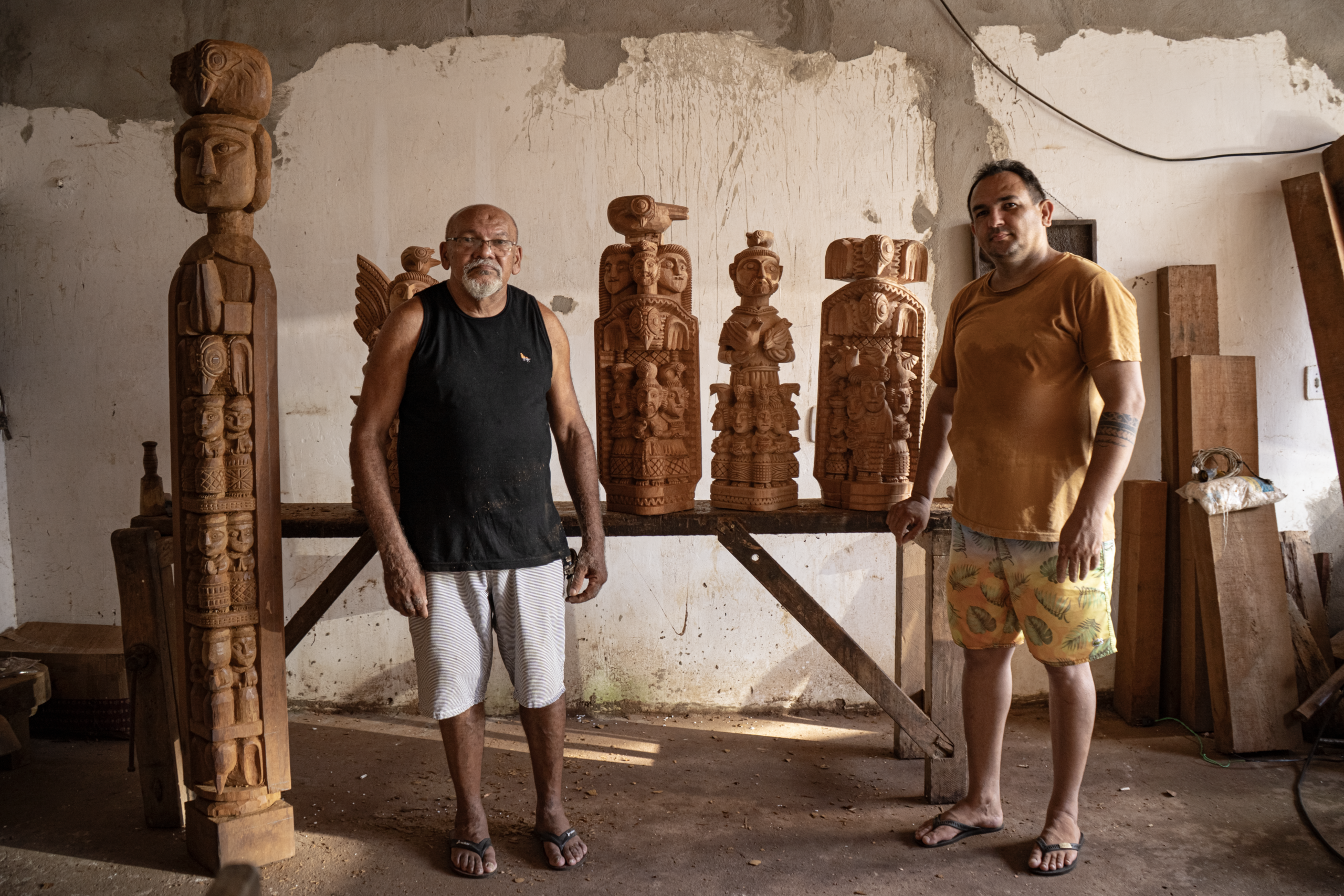 Dois artesãos posam ao lado de esculturas em madeira produzidas no ateliê, onde realizam trabalhos de entalhe.
