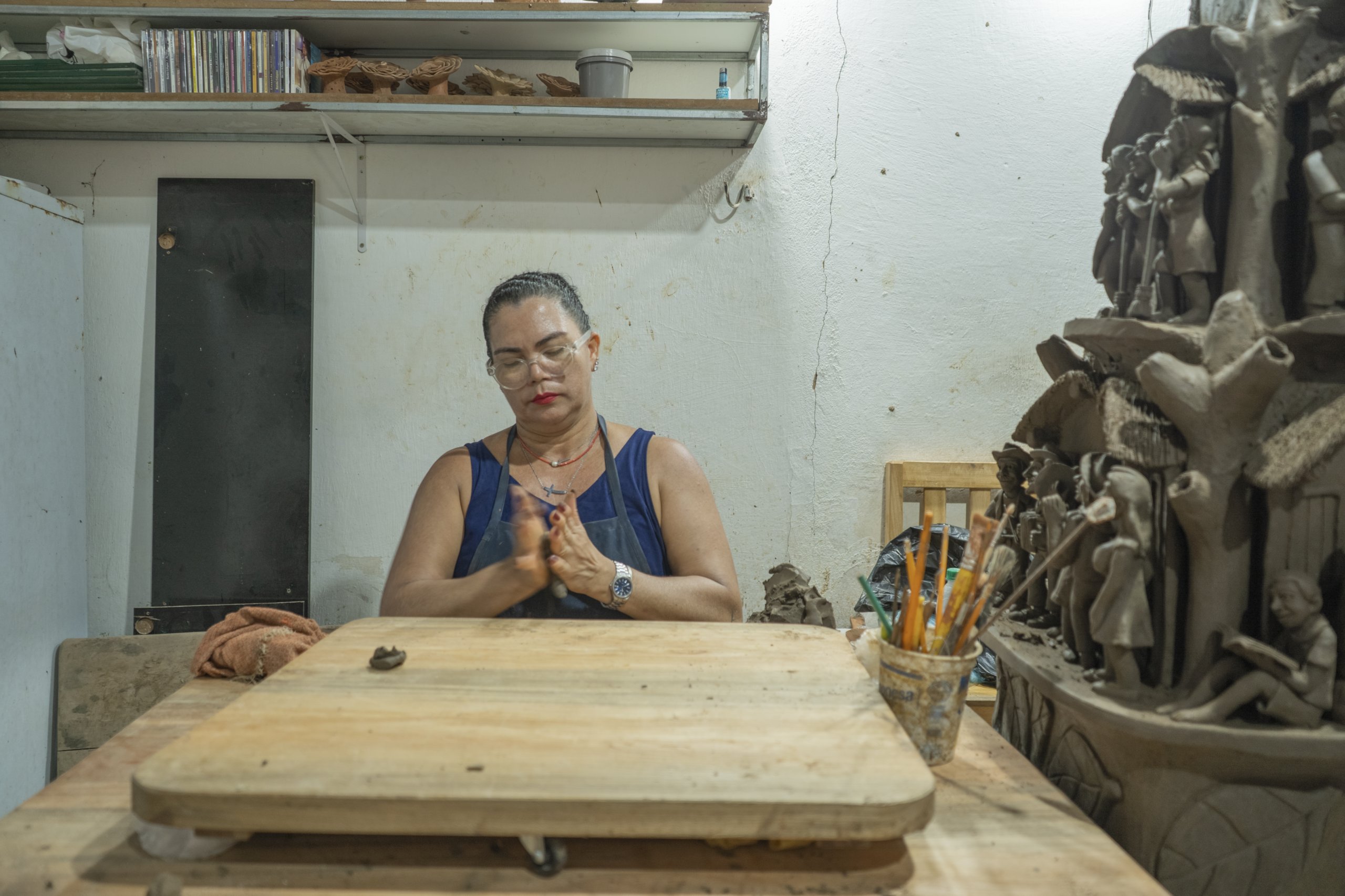 Artesã trabalha a argila sobre a mesa de produção em seu espaço de criação, cercada por ferramentas e esculturas em cerâmica.
